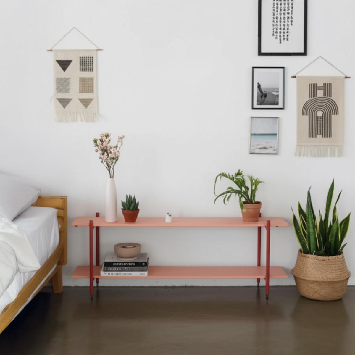 A long, low two-tier coral shelf beside a wooden bed, with a vase of flowers and two potted plants on top and a stack of books and a bowl on the lower shelf.