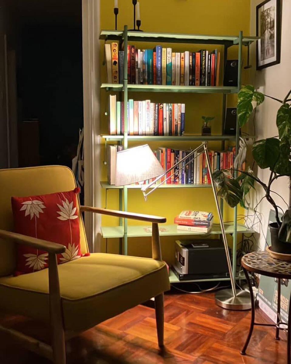 A tall sage shelf full of books against a yellow wall, with a reading lamp and a mustard armchair in front of it.