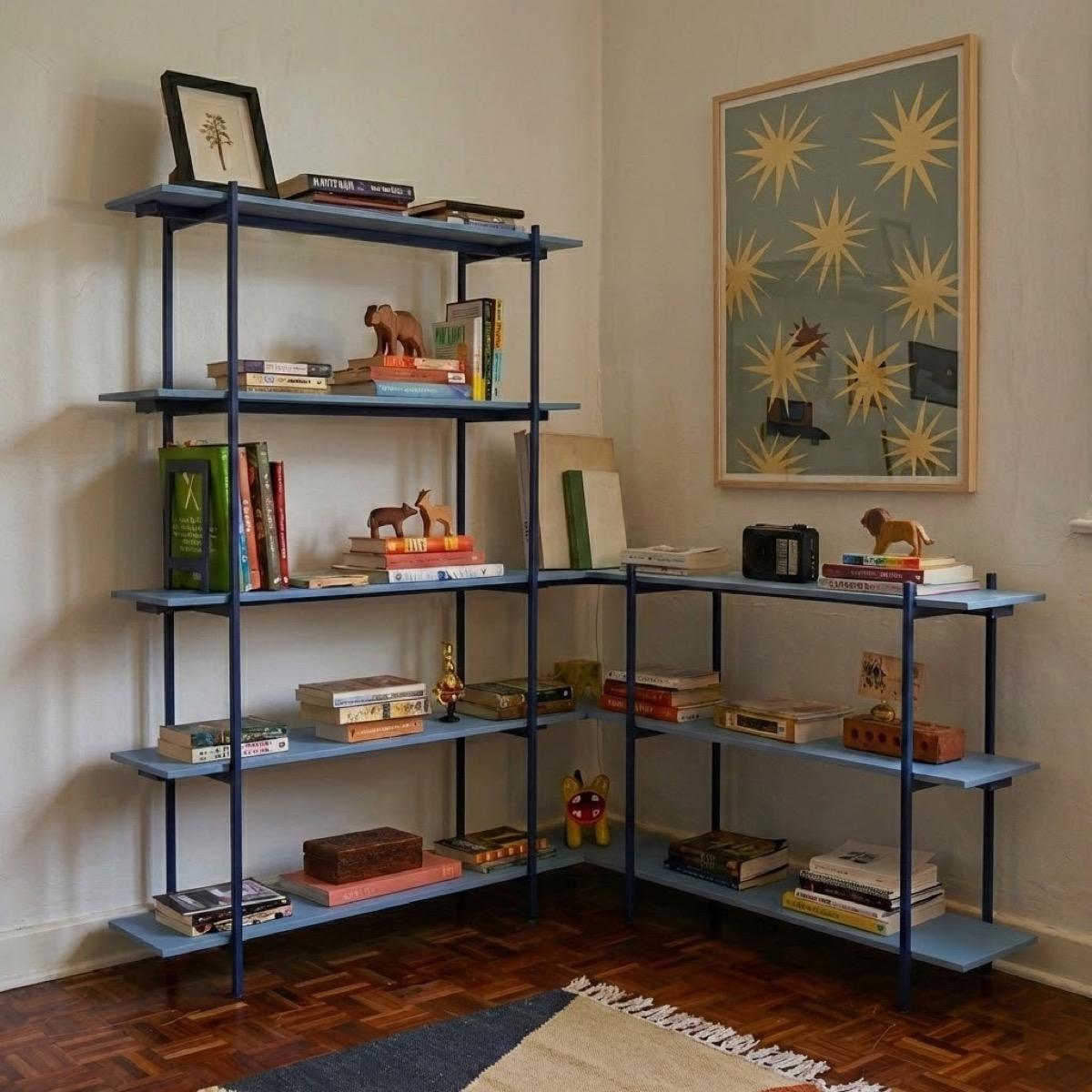 An L-shaped shelf in marine blue steel with pale blue boards in the corner of a room, taller on one wall and lower on the other, holding books, a small radio, carved wooden animals and a framed print, on a parquet floor.