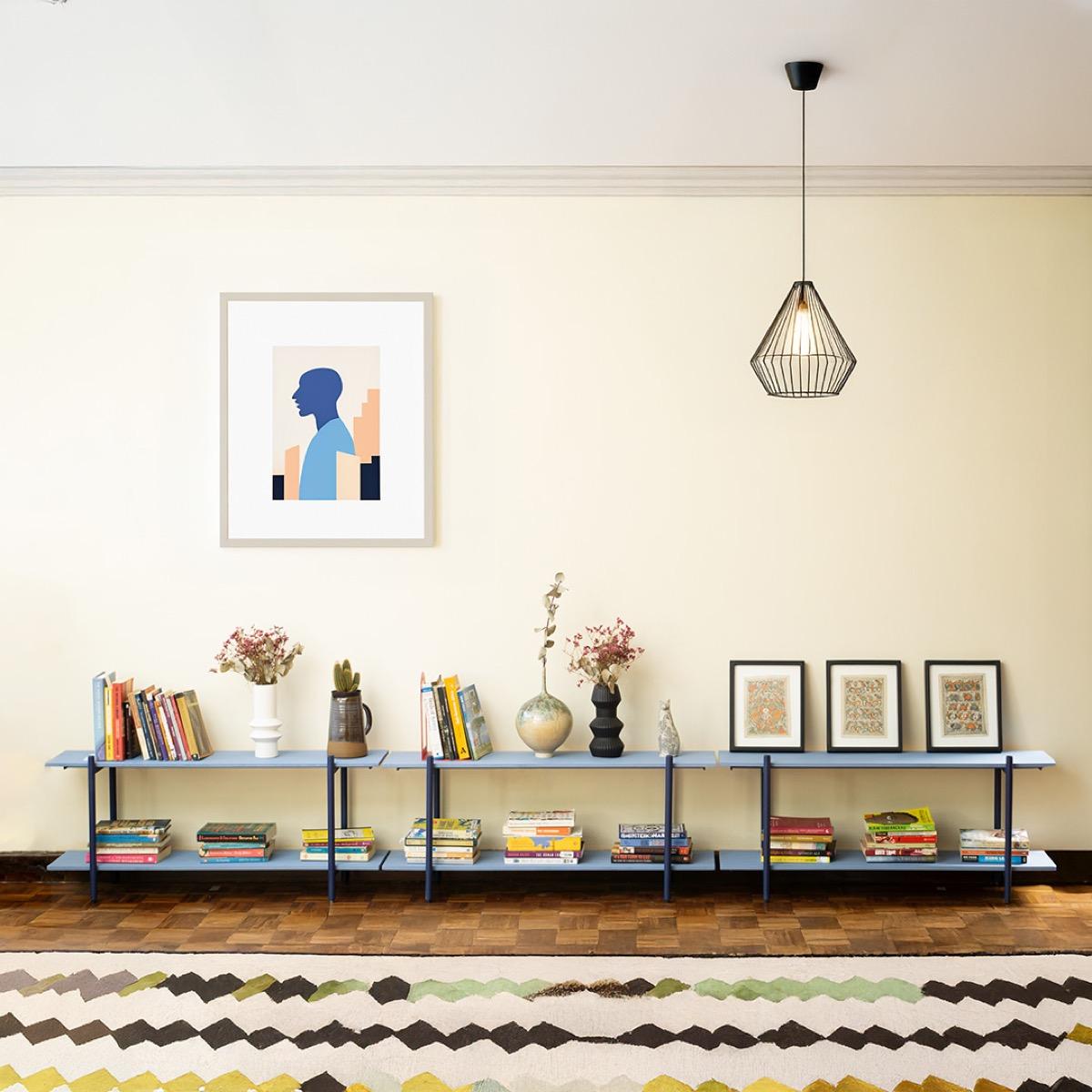 Three low two-tier shelves in a row along a cream living-room wall, with deep blue frames and pale blue boards, holding books, vases of dried flowers, a small cactus and three small framed prints, under a framed print and a wire pendant lamp.