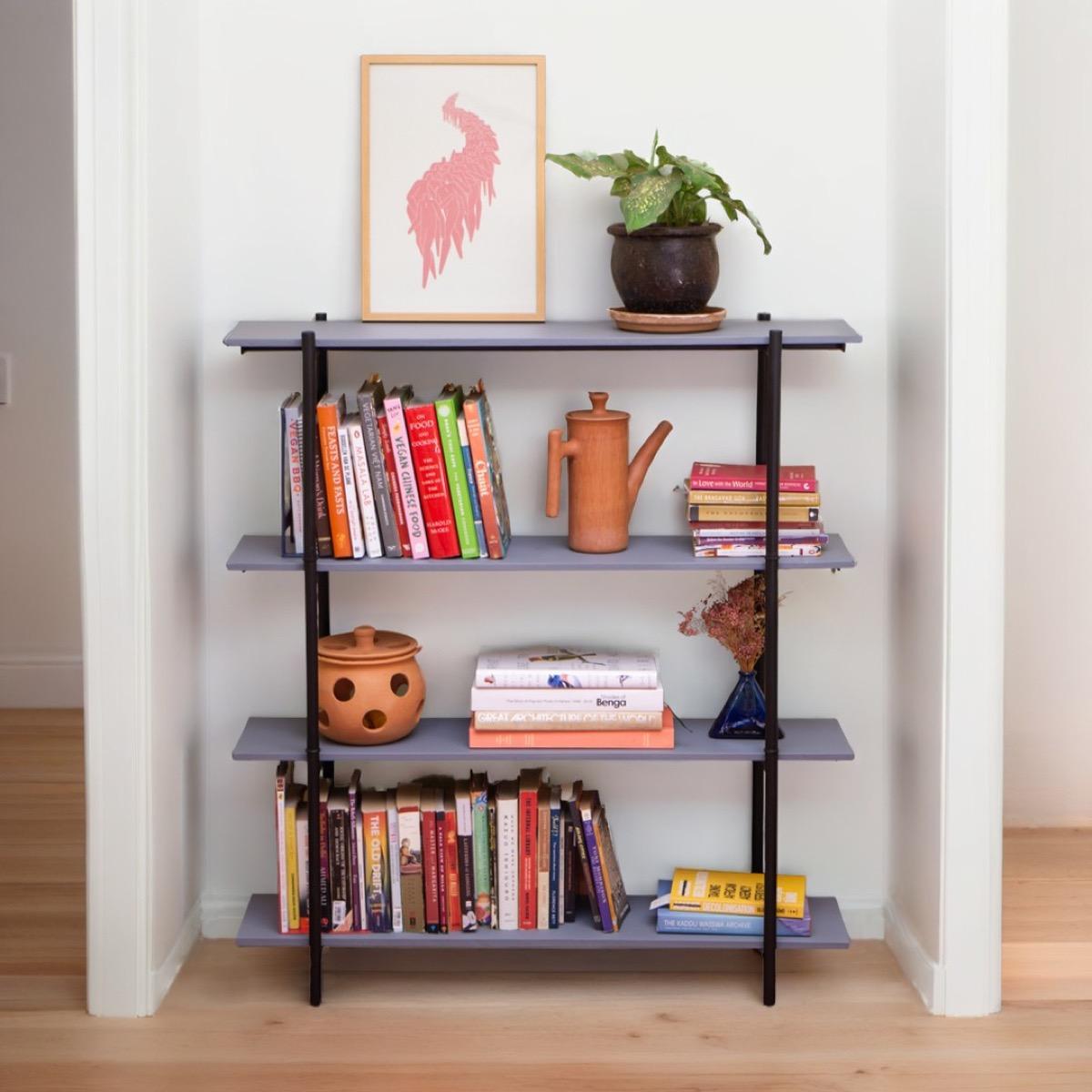 A four-shelf unit with charcoal steel legs and grey boards standing in a white alcove, holding rows of cookbooks and novels, a clay jug, a small vase of dried flowers, a potted plant and a framed pink print.