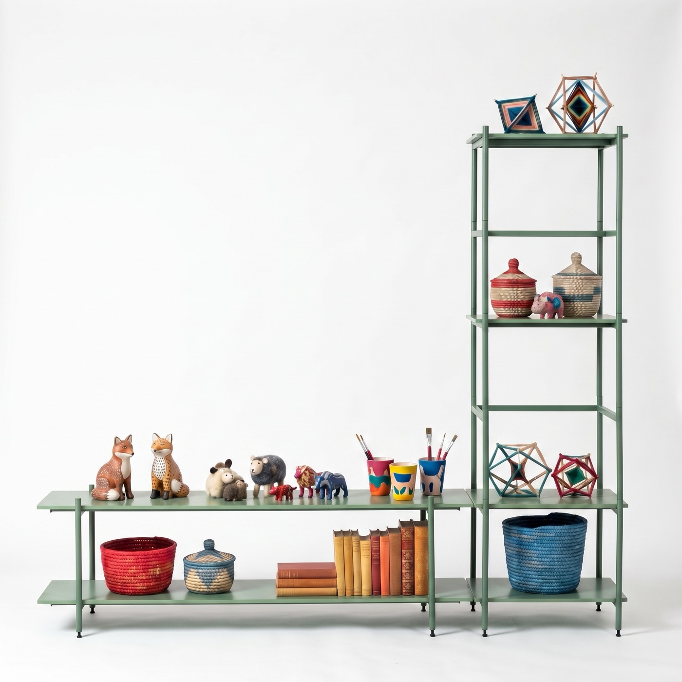 A sage green steel shelf in two parts: a long low unit with two shelves holding carved foxes, felt sheep, painted cups of brushes, woven baskets and old books, and a tall narrow tower beside it with lidded baskets and woven ornaments.