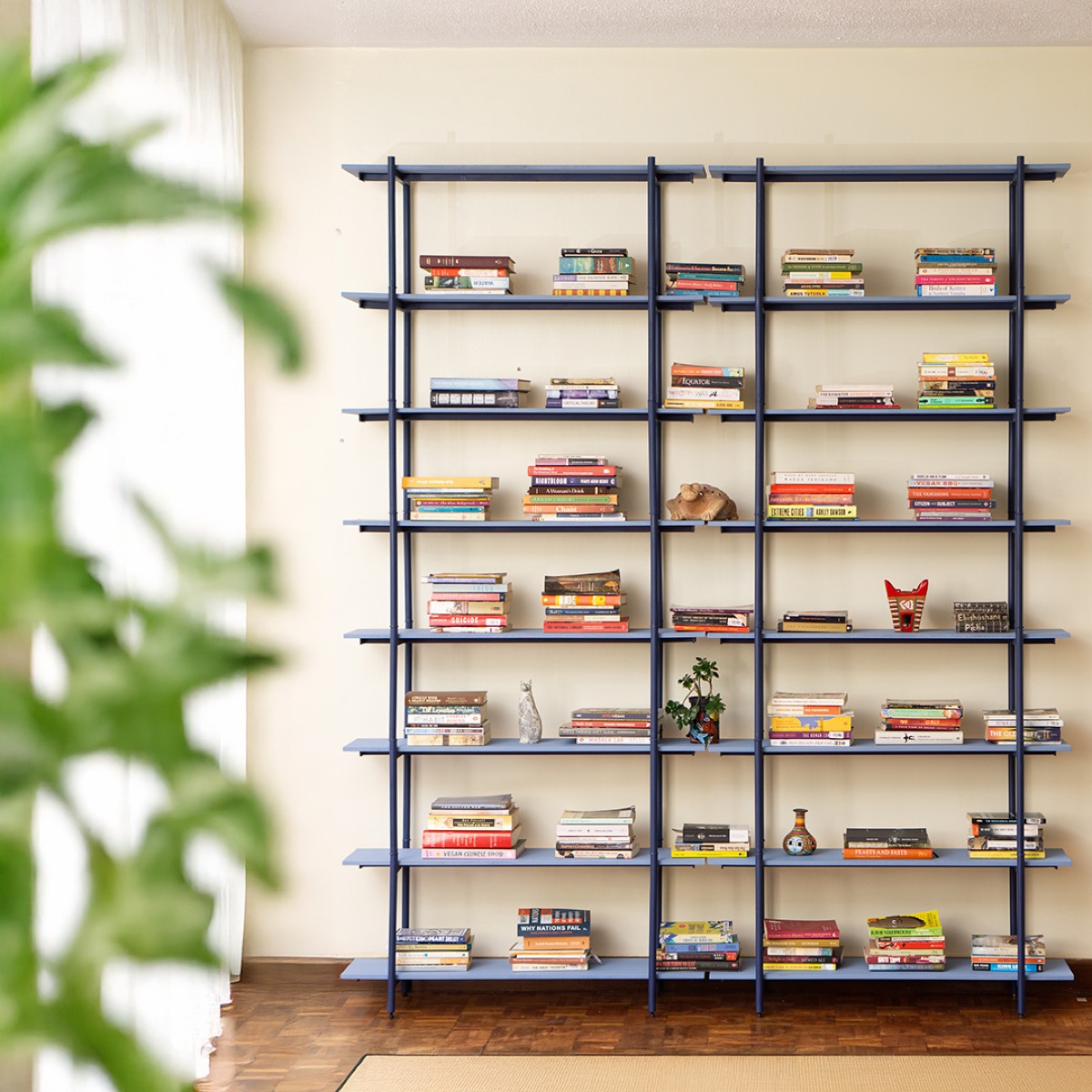 Two tall bays of shelving in marine blue steel against a cream wall, eight shelves high, with stacks of books on every shelf and a small bonsai, a vase and carved animals among them.