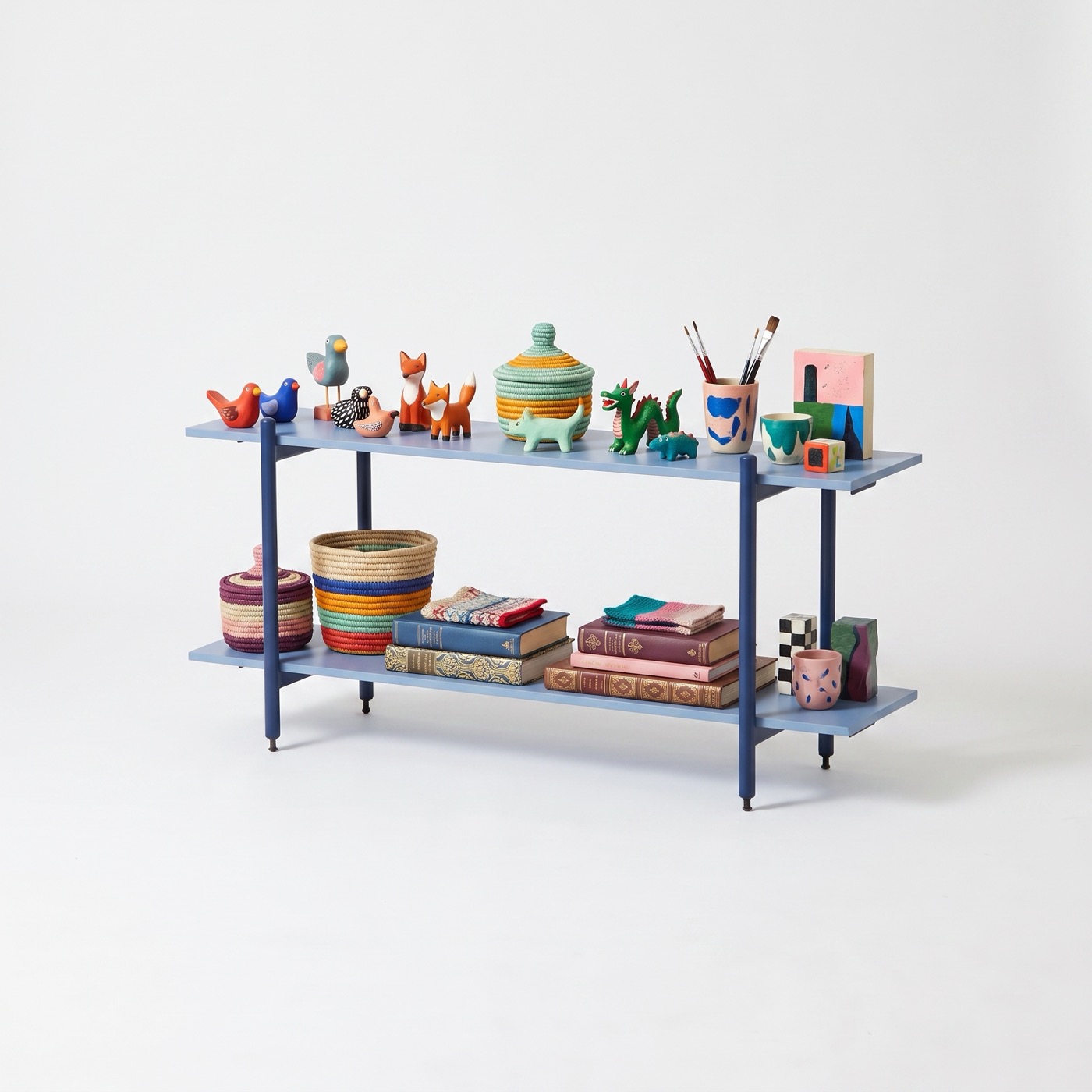 A low two-tier shelf in deep blue steel with pale blue boards, holding carved wooden birds and foxes, a lidded basket, paintbrushes in a cup, woven baskets and a stack of books.
