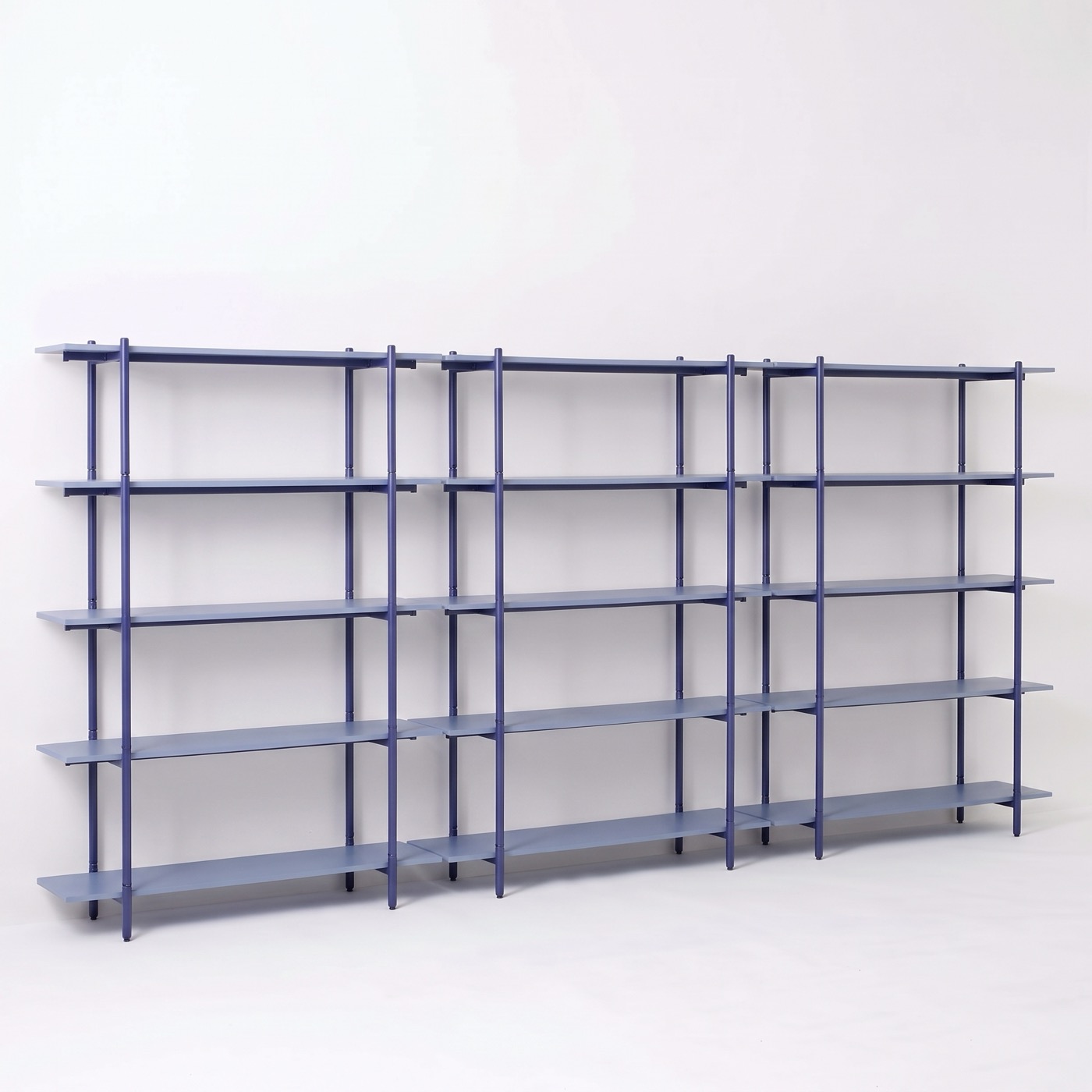 An empty three-bay shelf in marine blue steel, seen from an angle, five shelves in each bay, with the wall visible through the open back.
