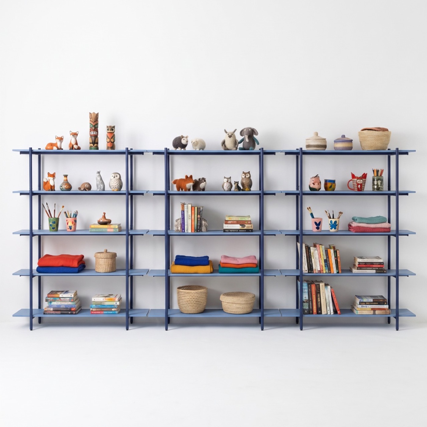 Three tall bays of marine blue steel shelving side by side, five shelves each, holding books, folded blankets, lidded baskets, pots of pencils and carved animals.